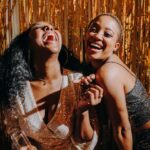 Two women laughing and enjoying a party with a festive golden backdrop.